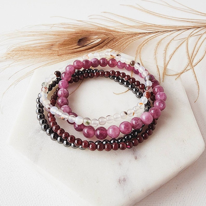 May include: A set of four bracelets made with different types of beads. The bracelets are layered on top of each other and feature pink, white, and black beads.