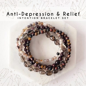 May include: Set of four bracelets made with brown, red, and gray gemstone beads. The bracelets are stacked on a white surface and the text "Anti-Depression & Relief Intention Bracelet Set" is displayed above the bracelets.