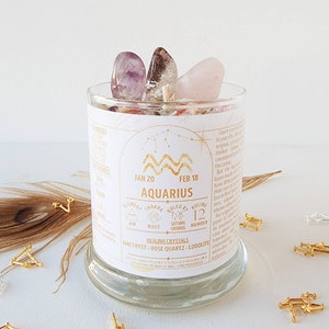 May include: A white candle jar with a gold label featuring the zodiac sign Aquarius, dates January 20 to February 18, and the text "Ruled by Saturn & Uranus". The jar contains amethyst, rose quartz, and lodalite crystals.