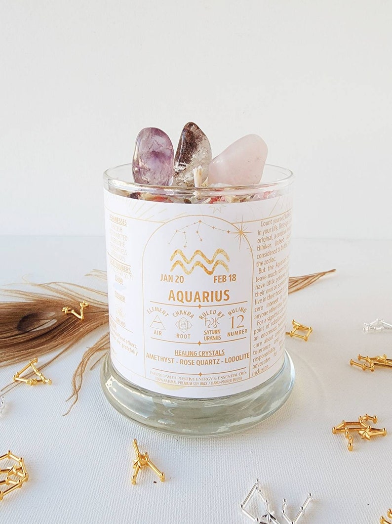 May include: A white candle jar with a gold label featuring the zodiac sign Aquarius, dates January 20 to February 18, and the text "Ruled by Saturn & Uranus". The jar contains amethyst, rose quartz, and lodalite crystals.