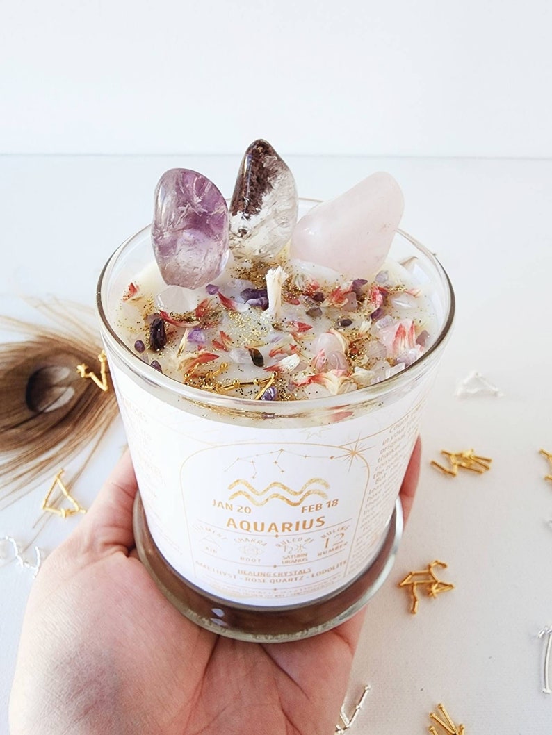 May include: A white candle with a clear glass jar and a gold label that says "Aquarius" with the dates "Jan 20" and "Feb 18". The candle is decorated with crystals, including amethyst, rose quartz, and clear quartz. The candle is surrounded by gold and silver decorative accents.