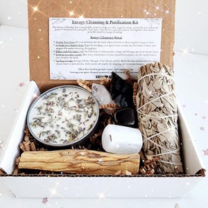 May include: A white box filled with items for an energy cleansing ritual. The box contains a white sage smudge stick, a palo santo stick, a lavender candle, a clear quartz crystal, a black obsidian arrowhead, and a selenite crystal. The box is labeled "Energy Cleansing & Purification Kit".