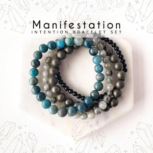 May include: Set of four beaded bracelets with blue, gray, and gold beads. The bracelets are stacked on a white surface and the text "Manifestation Intention Bracelet Set" is above the bracelets.