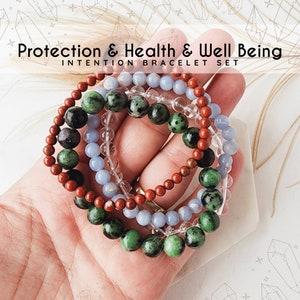 May include: Set of four bracelets made with green, red, blue, and clear beads. The bracelets are stacked on top of each other and say "Protection & Health & Well Being Intention Bracelet Set".