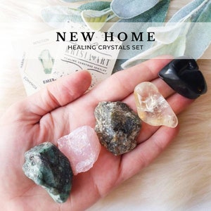 May include: A set of healing crystals for a new home. The set includes a black heart-shaped crystal, a clear crystal, a green crystal, a pink crystal, and a grey crystal. The crystals are arranged in a hand.