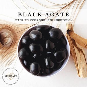 May include: A white bowl filled with black agate tumbled stones. The bowl is sitting on a white surface with a brown wooden branch in the background. The text "BLACK AGATE" and "STABILITY I INNER STRENGTH I PROTECTION" is displayed above the bowl.