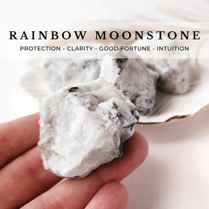 May include: A close-up of a white rainbow moonstone crystal with black inclusions. The crystal is being held in a hand. The text "RAINBOW MOONSTONE" and "PROTECTION - CLARITY - GOOD FORTUNE - INTUITION" is visible above the crystal.