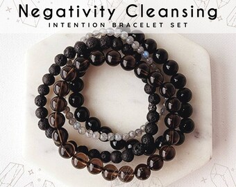 May include: Three stacked bracelets made of dark brown, black, and gray beads. The bracelets are on a white surface and the text "Negativity Cleansing Intention Bracelet Set" is above the bracelets.