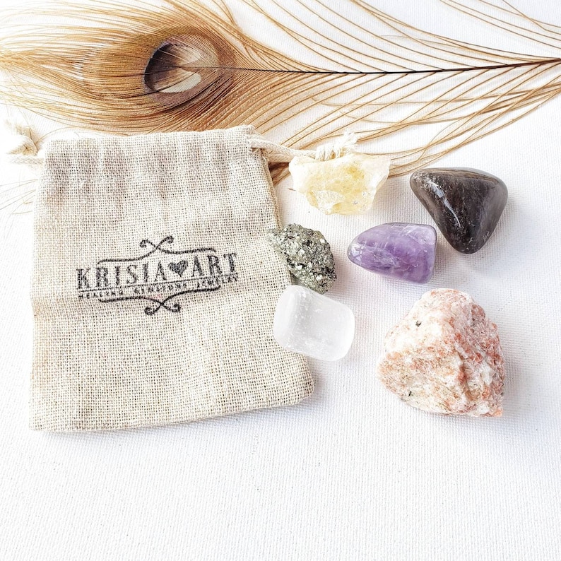 May include: A small, beige cloth bag with the text "Krisia Art" printed on it. The bag is surrounded by six different colored crystals: clear, yellow, purple, brown, green, and pink.
