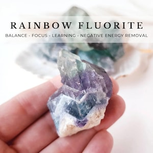 May include: A close-up of a rainbow fluorite crystal, showing its vibrant green, purple, and blue hues. The crystal is held in a hand, showcasing its natural shape and texture.  BALANCE - FOCUS - LEARNING - NEGATIVE ENERGY REMOVAL