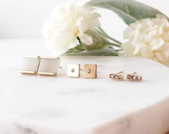 May include: Three pairs of gold earrings with different styles. The first pair features white square stones, the second pair has small gold squares with a single diamond in each, and the third pair is a thin gold bar with three small diamonds.