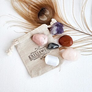 May include: A small pouch with a drawstring closure containing six tumbled stones: a pink and white stone, a dark gray stone, a purple stone, a red stone, and a white stone. The pouch is made of natural fabric and has the text "KRISIA LOVE" printed on it.