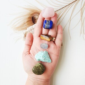 May include: A collection of seven crystals, including rose quartz, lapis lazuli, tiger's eye, clear quartz, green aventurine, and moss agate, arranged in a vertical line on a hand.