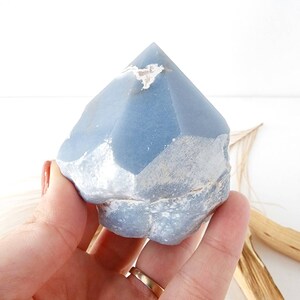 May include: A blue crystal point with a rough, natural texture. The crystal is a light blue color with white inclusions. It is a natural crystal and has a unique shape.