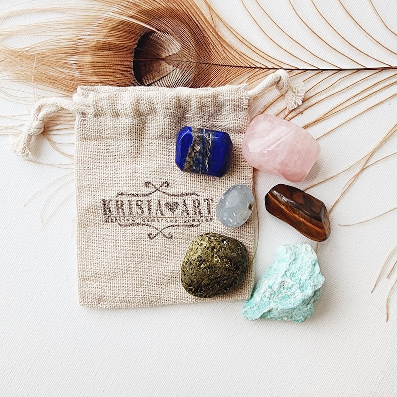 May include: A small pouch containing six tumbled crystals: blue lapis lazuli, pink rose quartz, clear quartz, brown tiger's eye, green aventurine, and turquoise. The pouch is made of natural fabric and has the text "KRISLA ART" printed on it.