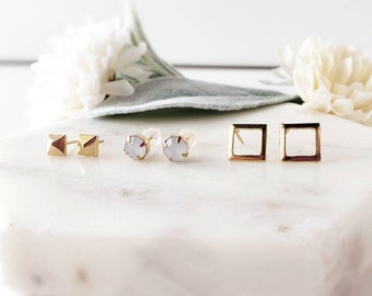 May include: Three pairs of gold stud earrings. The first pair is a geometric design with a pointed top. The second pair is a round, clear stone. The third pair is a square with an open center.