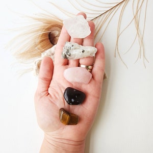 May include: A collection of five crystals, including clear quartz, rose quartz, tiger's eye, black obsidian, and a white crystal. The crystals are arranged in a row on a hand.