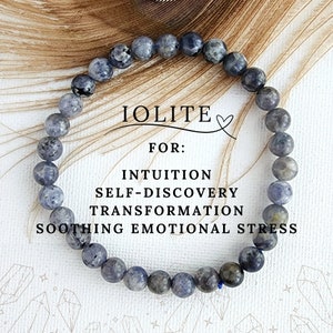 May include: A blue and grey iolite gemstone bracelet. The bracelet is made of round beads and is strung on elastic cord. The text "IOLITE" and "FOR: INTUITION SELF-DISCOVERY TRANSFORMATION SOOTHING EMOTIONAL STRESS" is written on a white background behind the bracelet.