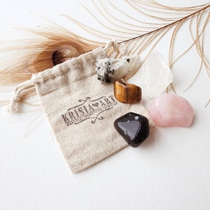 May include: A small, beige cloth bag with a drawstring closure, containing five tumbled crystals: clear quartz, tiger's eye, black obsidian, rose quartz, and a white crystal with black flecks. The bag has a brown logo that reads "Kristal Art".