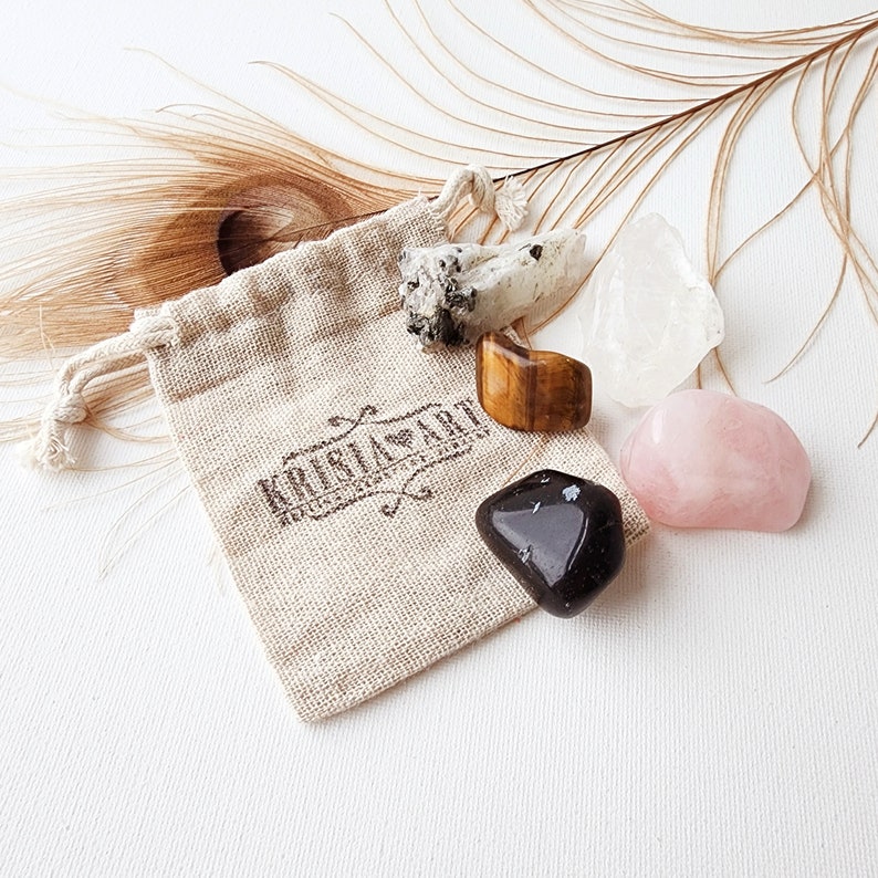 May include: A small, beige cloth bag with a drawstring closure, containing five tumbled crystals: clear quartz, tiger's eye, black obsidian, rose quartz, and a white crystal with black flecks. The bag has a brown logo that reads "Kristal Art".