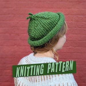 May include: A green knitted hat with a pointed top and a brim. The hat is being worn by a person with long brown hair. The text "Knitting Pattern" is visible in the image.