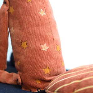 May include: A plush toy with a brown corduroy body and yellow star pattern. The toy has a long tail and a striped pillow.