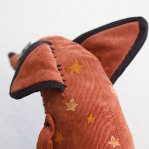 May include: A plush toy shaped like a fox with brown corduroy fabric and black trim. The fabric is patterned with gold stars.