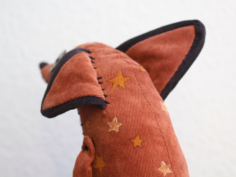 May include: A plush toy shaped like a fox with brown corduroy fabric and black trim. The fabric is patterned with gold stars.
