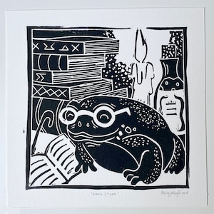 May include: Black and white linocut print of a frog wearing glasses and reading a book. The frog is sitting on a pile of books. The print is titled "Frank | Toad" and signed "Holly Glasgow 2019".