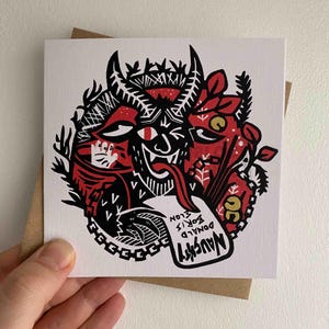 May include: A square greeting card featuring a red and black devil illustration. The devil has horns and a long tongue, with the word "Naughty" on a tag. The names "Donald, Boris, Elon" are also on the tag. The card is on a brown envelope.