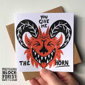 Peut inclure: Carte de vœux carrée avec une illustration de démon cornu rouge et noir et le texte "YOU GIVE ME THE HORN". La carte est tenue au-dessus d'une enveloppe marron. Le logo Block Forest est dans le coin inférieur gauche.