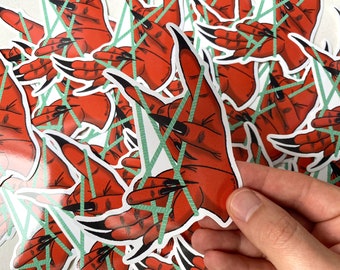 May include: A stack of red hand stickers with long black nails and green string wrapped around the fingers. The stickers are on a white surface.