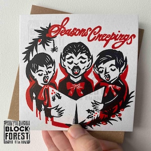 May include: A square greeting card with a brown envelope. The card features a black and red illustration of three vampire carolers singing from a book. The text "Seasons Creepings" is written in red script above the vampires.