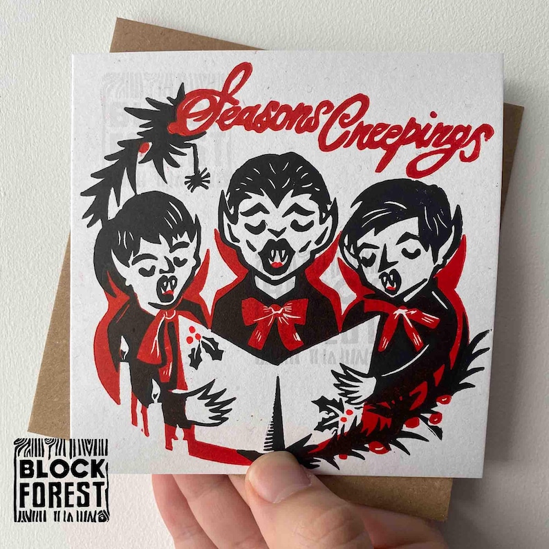 May include: A square greeting card with a brown envelope. The card features a black and red illustration of three vampire carolers singing from a book. The text "Seasons Creepings" is written in red script above the vampires.