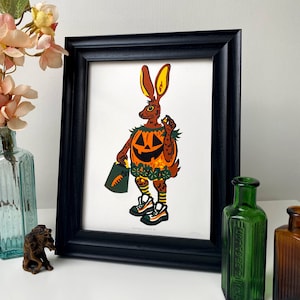Peut inclure: Une illustration encadrée d'un lapin déguisé en citrouille d'Halloween. Le lapin porte un costume vert et orange avec un visage de citrouille sculpté. Le lapin tient un petit seau orange avec une carotte à l'intérieur.