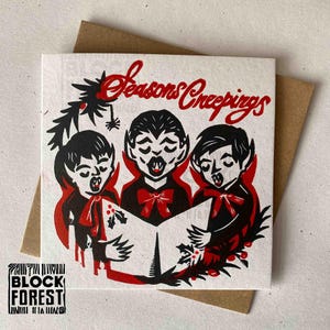 May include: A square greeting card with a brown envelope. The card features a red and black illustration of three vampire children singing carols, with the words "Seasons Creepings" in red script. The card has a logo in the bottom left corner.
