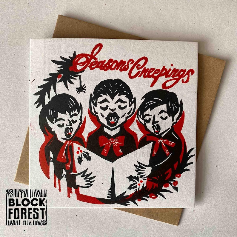 May include: A square greeting card with a brown envelope. The card features a red and black illustration of three vampire children singing carols, with the words "Seasons Creepings" in red script. The card has a logo in the bottom left corner.