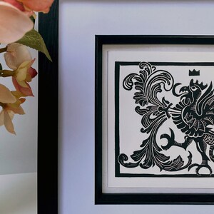May include: Black and white woodblock print of a crowned griffin with a floral design in a black frame.