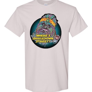 May include: A white t-shirt with a colorful graphic of a furry creature wearing a headband with the text "Where's Shakedown Street?" printed on the front.