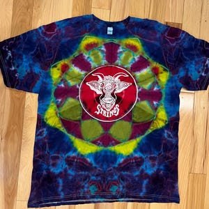 May include: A tie-dye t-shirt with a red circle and a white goat with one eye in the center. The circle has the text "Strings" in white.