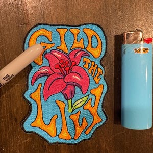 May include: Embroidered patch with the words "GILD THE LILY" in orange lettering on a blue background. The patch features a pink flower with green leaves. A blue BIC lighter is next to the patch.