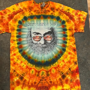 May include: A tie-dye t-shirt with a portrait of Jerry Garcia and the text "Jerry Garcia Band." The shirt features a vibrant spiral design in shades of orange, yellow, blue, and green. The graphic is centered on the chest.