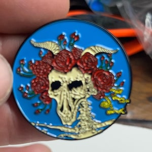 May include: A blue enamel pin with a skeleton wearing a crown of red roses. The skeleton has horns and is on a blue background.