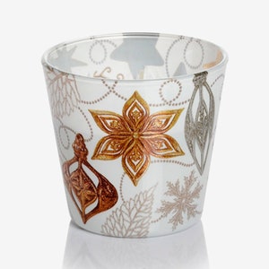 May include: A white glass candle holder with a festive design featuring gold, silver, and copper ornaments and snowflakes.