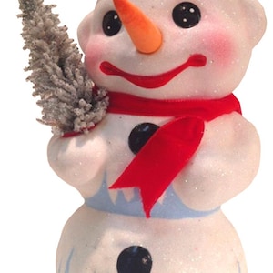 May include: A whimsical snowman figurine with a black top hat, orange carrot nose, and red scarf. The snowman holds a small, snow-covered tree. The base and bottom section of the snowman are light blue with white accents. The figurine is covered in glitter.