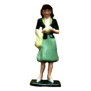 May include: A ceramic figurine of a woman in a green skirt and jacket, holding a yellow scarf. The figurine has brown hair and is standing on a black base. The jacket has black buttons and a yellow collar.
