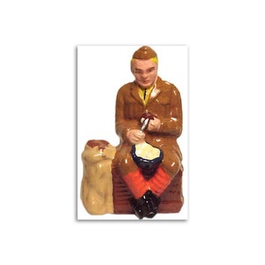 May include: A painted figurine of a soldier wearing a brown uniform and a yellow hat, sitting on a brown bench with a brown bag beside him. He is holding a spoon and eating from a bowl.