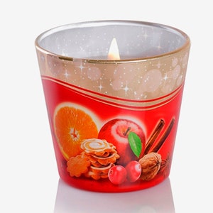 May include: A glass candle jar with a red, gold, and white design. The jar features a festive design with an orange slice, apple, cinnamon sticks, cranberries, and a walnut. The candle is lit and the flame is visible.