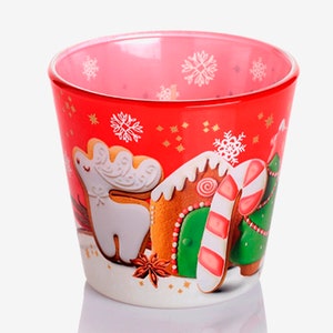 May include: Red glass mug with a Christmas theme. The mug features a gingerbread house, a candy cane, a Christmas tree, and a reindeer, all made of gingerbread. The mug is decorated with snowflakes and stars.