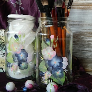 Puede incluir: Dos tarros de vidrio con diseños florales pintados, llenos de pinceles de maquillaje. Los tarros están sobre una tela morada con canicas blancas y azules.